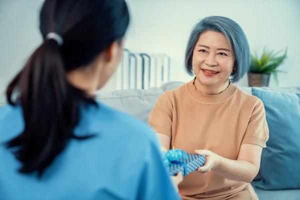 stock image A young caregiver hand over to her senior patient a blue gift box with blue ribbons at a contented living room.