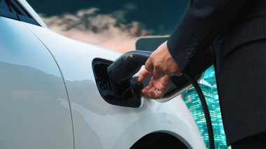 İlerici işadamı elektrikli aracını şarj kablosu ile şarj ediyor. Şarj istasyonundan şehrin arka planına bakıyor. Modern temiz enerji kampanyası konsepti için..