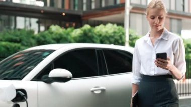 Şehir merkezinde dolaşmadan önce elektrikli araba ve şarj istasyonuna yaslanan ilerici iş kadını. Çevre dostu şarj edilebilir EV araba sürdürülebilir ve temiz enerjiyle çalışıyor.