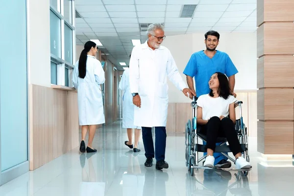 stock image Doctor and male nurse transport a female patient in a wheelchair along sterile hospital corridor. Health care and nursing care for disabled handicapped patient in the hospital concept.