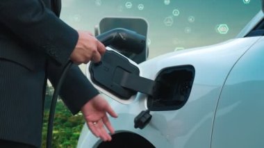 İlerleyen yeşil şehir ESG sembolü, elektrikli araçla birlikte. İş adamı EV arabalarını şarj etmek için şarj aletini şarj ediyor. Şehirdeki şarj istasyonunda ekolojik ve yeşil semboller var..