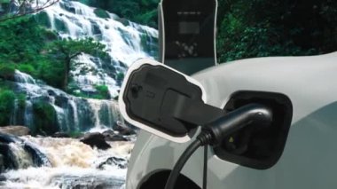 Elektrikli araba, doğal ortam, şelale ve akış arkaplanındaki şarj istasyonundan enerji yüklüyor. Yenilenebilir enerji kaynaklı EV otomobillerin enerji sürdürülebilirliği kavramı.