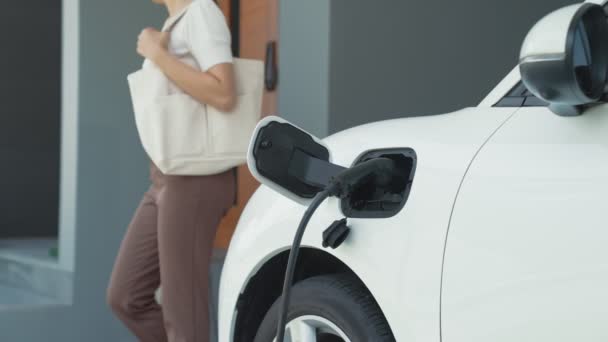 女性が電気自動車の充電器を彼の家に取り外します 先進的なライフスタイルにおける電気自動車の使用の概念は 清潔で健康的な環境に貢献します — ストック動画