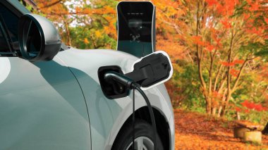 İlerici çevre dostu elektrikli araba konsepti, temiz ve sürdürülebilir enerji şarj istasyonundan güç alan EV otomobili turuncu sonbahar ormanlarının arka planına karşı.