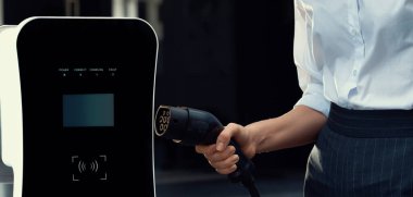 Focus EV charger plug and electric car at public charging station with blur progressive businesswoman holding charger and apartment condo building in background. Eco friendly electric vehicle concept.