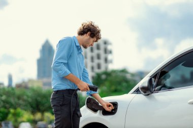 Progressive businessman insert charger plug from charging station to his electric vehicle with apartment condo building in background. Eco friendly rechargeable car powered by sustainable energy.
