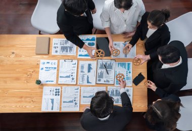 Top view hand holding gear by businesspeople wearing suit for harmony synergy in office workplace concept. Group of people hand making chain of gears into collective form with dashboard report papers.