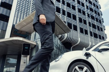 Closeup progressive businessman with electric car recharging at public charging station with background of city residential building. Eco friendly car powered by alternative clean energy.