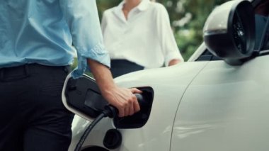İlerici işadamı ve iş kadını, şehir merkezinde dolaşmadan önce şarj cihazını şarj istasyonundan elektrikli arabaya takıyor. Çevre dostu şarj edilebilir araba temiz enerjiyle çalışıyor..