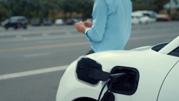電源ケーブルプラグと再生可能エネルギー車と充電ステーションに立っている間 彼の電気自動車にもたれて進歩的な野心を持つスーツを身に着けているビジネスマン — ストック動画