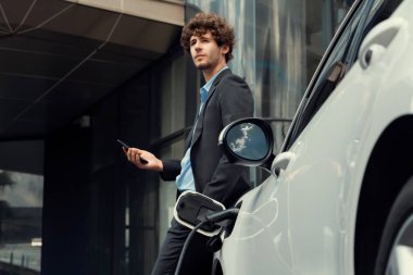 Progressive businessman talking on the phone, leaning on electric car recharging with public EV charging station, apartment condo residential building on the background as green city lifestyle.