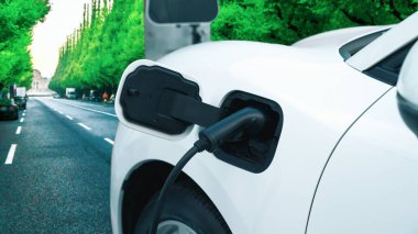 Çağdaş yeşil şehir ESG yeşil ağaçlar elektrikli araçla arka planda, EV araba şarj noktasındaki enerji yenilenebilir enerji taşımacılığıyla çevre bilincini artırmak için şarj ediliyor.