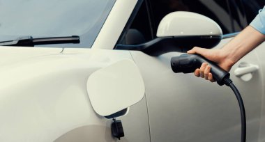 Closeup progressive man holding EV charger plug from public charging station for electric vehicle with background of residential building as concept eco-friendly sustainability energy car concept.