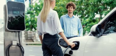 Progressive businessman and businesswoman with electric car parking and connected to public charging station before driving around city center. Eco friendly rechargeable car powered by clean energy.