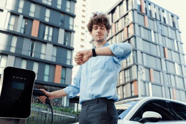 Businessman with smartwatch at modern charging station for electric vehicle with background of residential buildings as concept for progressive lifestyle of using eco-friendly as alternative energy.