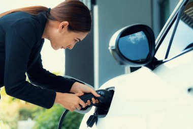 Progressive woman install cable plug to her electric car with home charging station. Concept of the use of electric vehicles in a progressive lifestyle contributes to clean environment.
