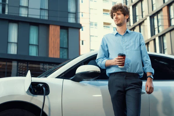 Progressive eco-friendly concept of parking EV car at public electric-powered charging station in city with blur background of businessman leaning on recharging-electric vehicle with coffee.