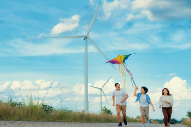 Progressive happy carefree family vacation concept. Young parents mother father and son run along and flying kite together on road with natural scenic on mountain and wind turbine background.