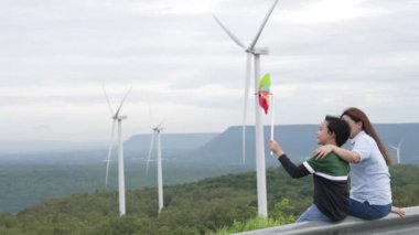 İlerici mutlu anne ve oğlu rüzgâr türbini çiftliğinde. Rüzgar türbini jeneratörüyle gelen elektrik jeneratörü kırsal bölgede tepe ve ufukta dağ var..