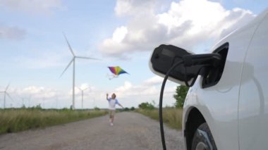 Rüzgar türbini çiftliğinde EV arabasının yanında yel değirmeni oyuncağı oynayan ilerici genç çocuk. Kırsal kesimdeki rüzgar türbini jeneratöründen elektrik jeneratörü..