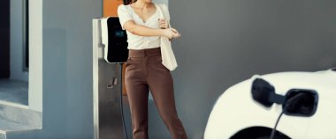 Focus EV charger plugged into EV car at home charging station with blurred background of progressive woman walking in background. Elective vehicle powered by clean energy for eco-friendly concept.