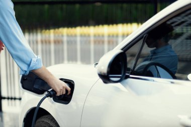 Closeup progressive man holding EV charger plug from public charging station for electric vehicle with background of residential building as concept eco-friendly sustainability energy car concept.