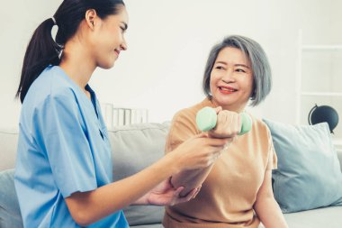 Caregiver helping contented senior woman exercise with dumbbell at home. Rehab for elder, senior medical care.