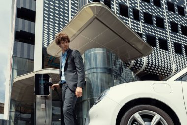 Below view of progressive businessman with electric car recharging at public charging station at modern city residential building. Eco friendly rechargeable EV car powered by alternative clean energy.