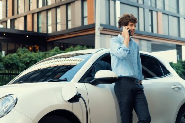 Progressive businessman talking on the phone, leaning on electric car recharging with public EV charging station, apartment condo residential building on the background as green city lifestyle.