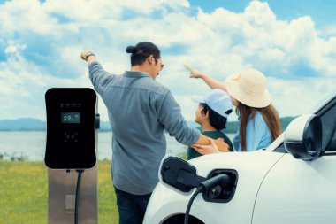 Concept of progressive happy family with electric vehicle enjoy their time at natural outdoor scenic with lake, greenfield and cloudscape background. Electric vehicle driven by green renewable energy.