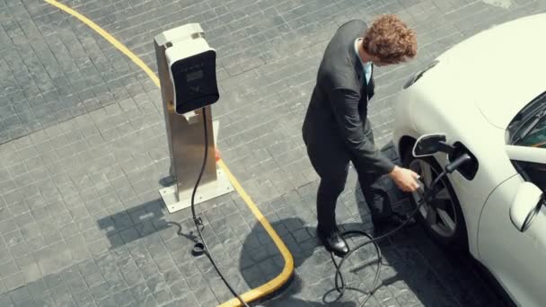 プログレッシブビジネスマンは 充電ステーションから彼の電気自動車に充電プラグを差し込み 市内中心部を走行します 持続可能でクリーンなエネルギーを動力とする環境に優しい充電式車 — ストック動画