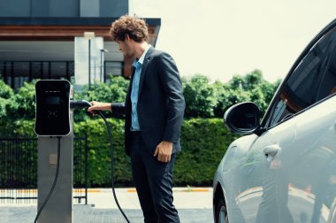 Modern şehir yerleşim bölgesindeki kamu şarj istasyonunda elektrikli araba şarj eden yenilikçi bir işadamı. Çevre dostu şarj edilebilir araba alternatif temiz enerjiyle çalışıyor..