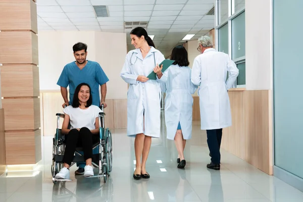 stock image Doctor and male nurse transport a female patient in a wheelchair along sterile hospital corridor. Health care and nursing care for disabled handicapped patient in the hospital concept.