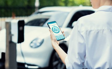 Suit-clad progressive businesswoman look at EV cars battery status from her phone while standing on a charging station with a power cable plug and a renewable energy-powered electric vehicle.