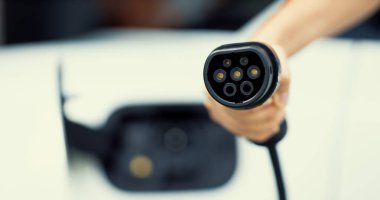 Progressive new alternative clean energy vehicle and electric charging device concept. Focus closeup hand holding and pointing EV charger plug at camear from electric charging station at camera