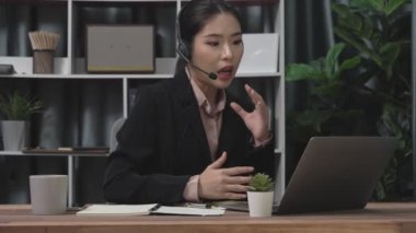 Kulaklık ve mikrofon takan iş kadını masasında çalışıyor ve dizüstü bilgisayar kullanıyor. Uzaktan kumandalı online toplantı için dizüstü bilgisayar kullanan hevesli kadın çalışanlar, ofiste iş videosu konferansı.