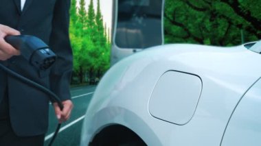 İlerici işadamı yeşil ormanın yakınındaki şarj noktasından elektrikli aracını şarj etmek için duruyor. Gelecekteki ortamı korumak için sürdürülebilir ve temiz enerji tarafından sürülen EV otomobil.