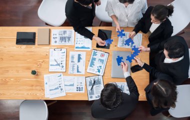 Top view businesspeople and colleagues in formal wear putting jigsaw puzzles together over meeting table with financial report papers in harmony office for team building concept.