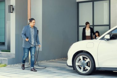 Progressive young parents and daughter with electric vehicle and home charging station. Green and clean energy from electric vehicles for healthy environment. Eco power from renewable source at home.