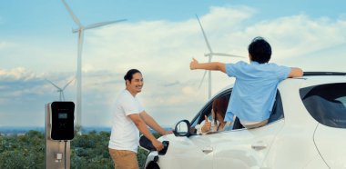 İlerici mutlu aile kavramı elektrikli araçla rüzgar çiftliğinde eğleniyor. Rüzgar türbini jeneratöründen gelen temiz yenilenebilir enerji ile şarj edilen elektrikli araç..