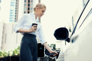 Businesswoman drinking coffee, leaning on electric vehicle recharging at public charging station with residential apartment condos building in background as progressive lifestyle by eco-friendly car.