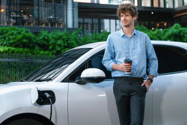 Progressive eco-friendly concept of parking EV car at public electric-powered charging station in city with blur background of businessman leaning on recharging-electric vehicle with coffee.