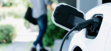 Focus electric car charging at home charging station with blurred progressive man walking in the background. Electric car using renewable clean for eco-friendly concept.