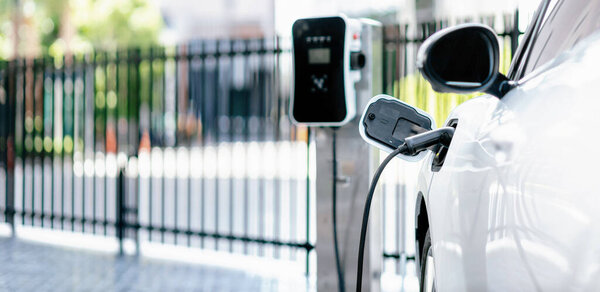 Focus closeup electric vehicle plugged in with EV charger device from blurred background of public charging station powered by renewable clean energy for progressive eco-friendly car concept.