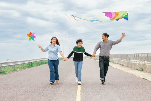 Progressive happy family vacation and carefree day concept. Young parents mother father and son run along and flying kite together road with enjoy natural scenic on scenery and clear sky background.