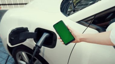 Taklit uzay için şarj istasyonuna bağlı elektrikli aracın yeşil ekran enerji durumunu gösteren bir model telefon. Temiz çevre için ilerici bir kavram. EV, yeşil yenilenebilir enerjiden güç alıyor.