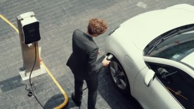 İlerici işadamı, şehir merkezinde dolaşmadan önce şarj cihazından elektrikli arabasına şarj prizi takıyor. Çevre dostu şarj edilebilir araba, sürdürülebilir ve temiz enerjiyle çalışıyor..