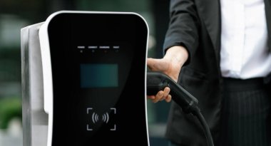 Focus EV charger plug and electric car at public charging station with blur progressive businesswoman holding charger and apartment condo building in background. Eco friendly electric vehicle concept.