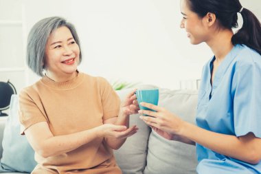 Female care taker serving her contented senior patient with a cup of coffee at home, smiling to each other. Medical care for pensioners, Home health care service.