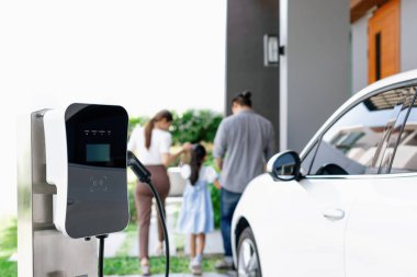 Focus closeup electric vehicle recharging battery from home electric charging station with blurred family in background. Renewable clean energy car for progressive eco awareness lifestyle concept.